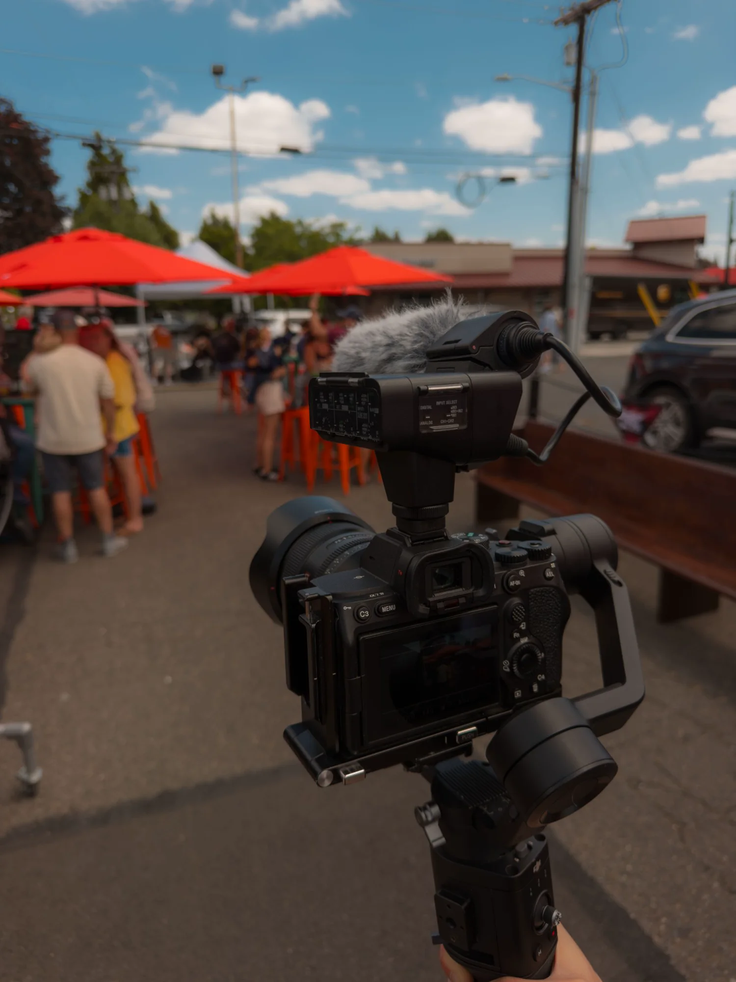 Sony camera with an XLR audio handle and deadcat mic on a DJI Ronin gimbal, filming a busy patio under red umbrellas on a summer day